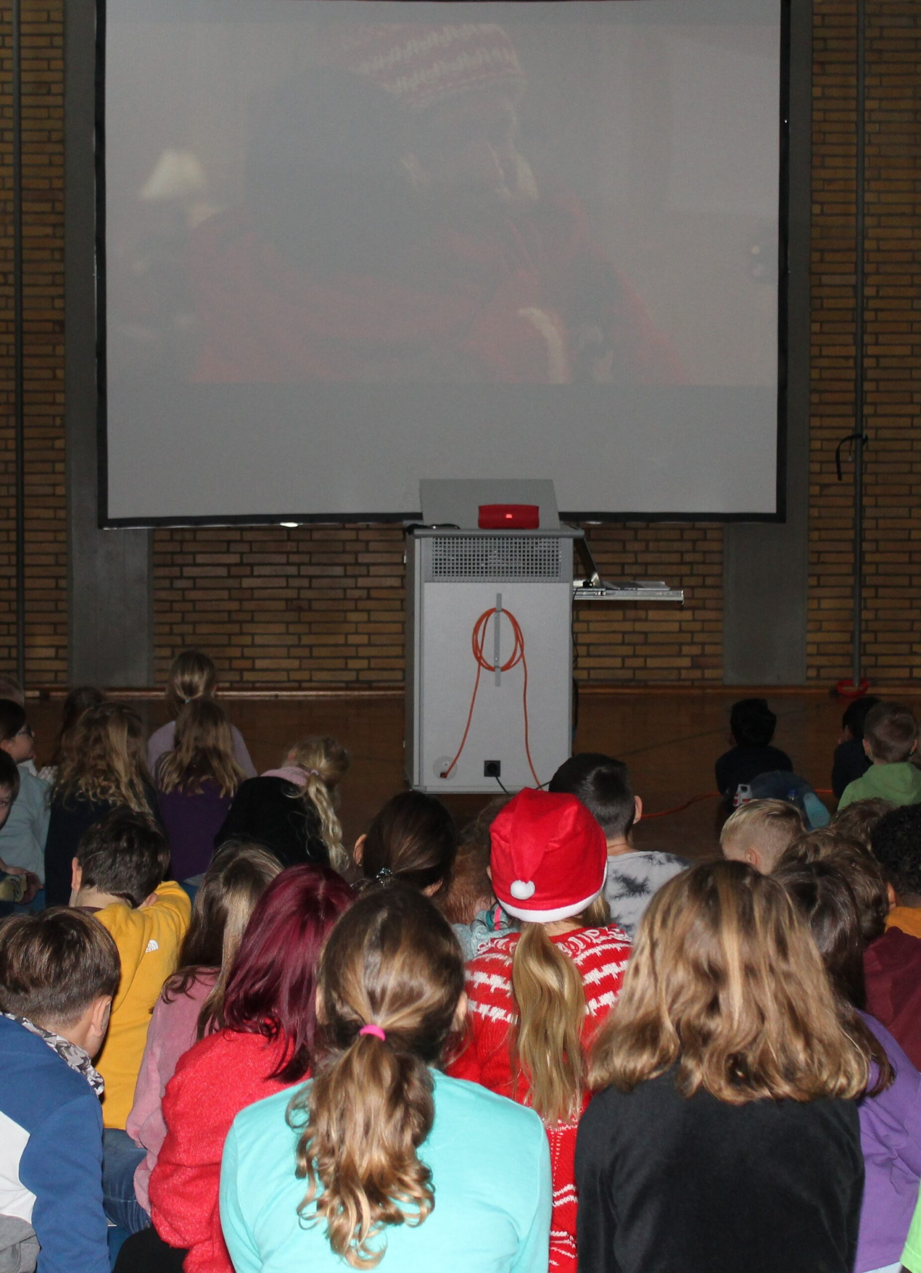 Schulkinder sitzen in der Sporthalle und schauen auf einer großen Leinwand einen Weihnachtskinofilm an.