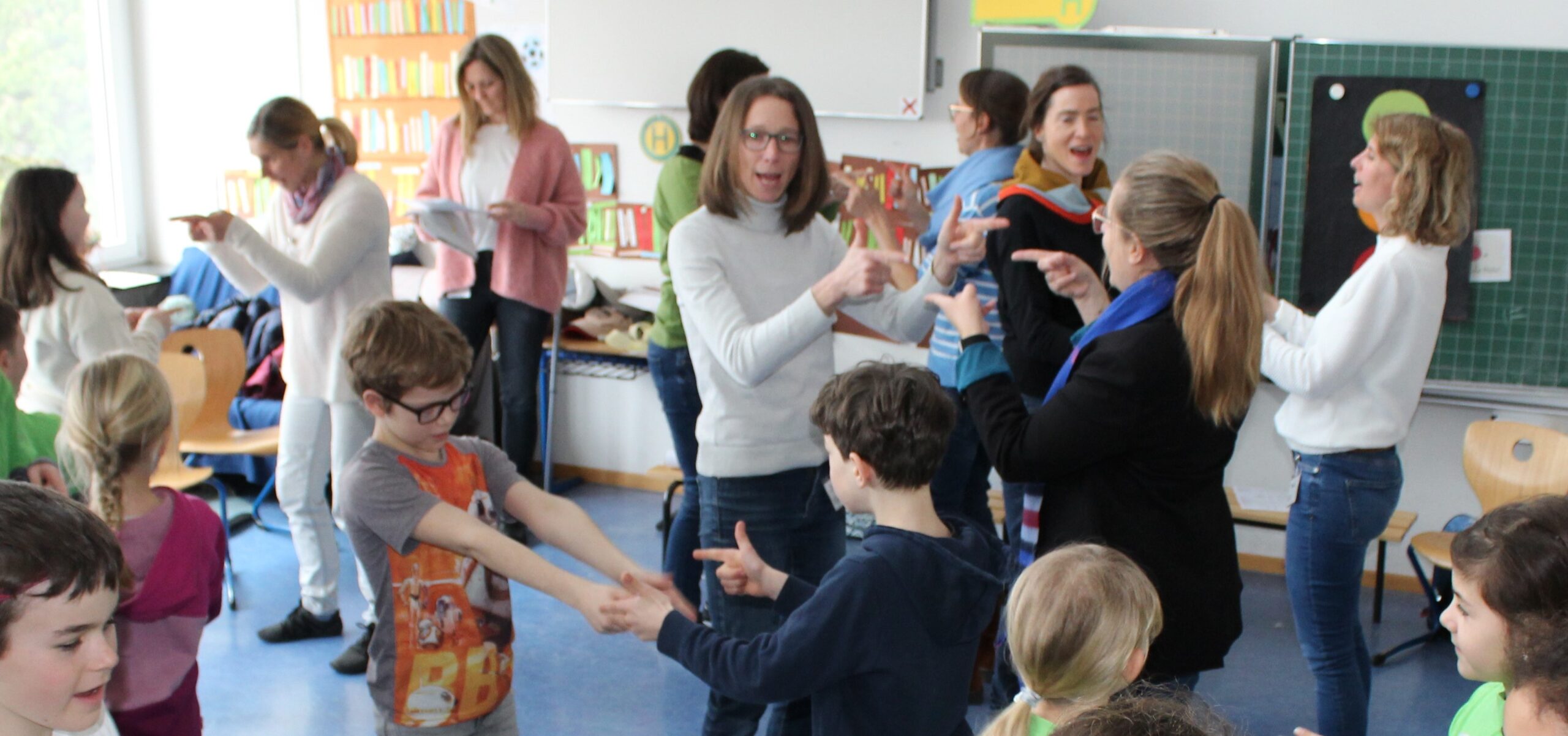 Eltern und Kinder tanzen in einem Klassenzimmer ein Freundschaftslied.
