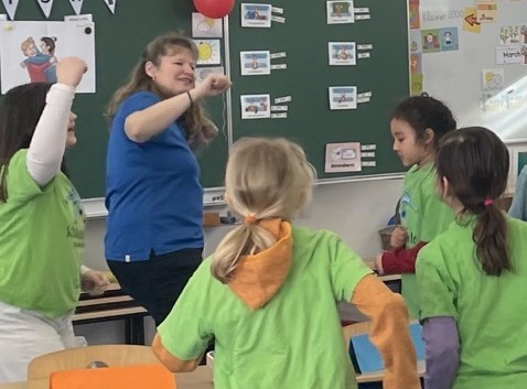 Eine Gesundheitsförderin macht zusammen mit Kindern einer Schulklasse Bewegungen zu einem Lied.