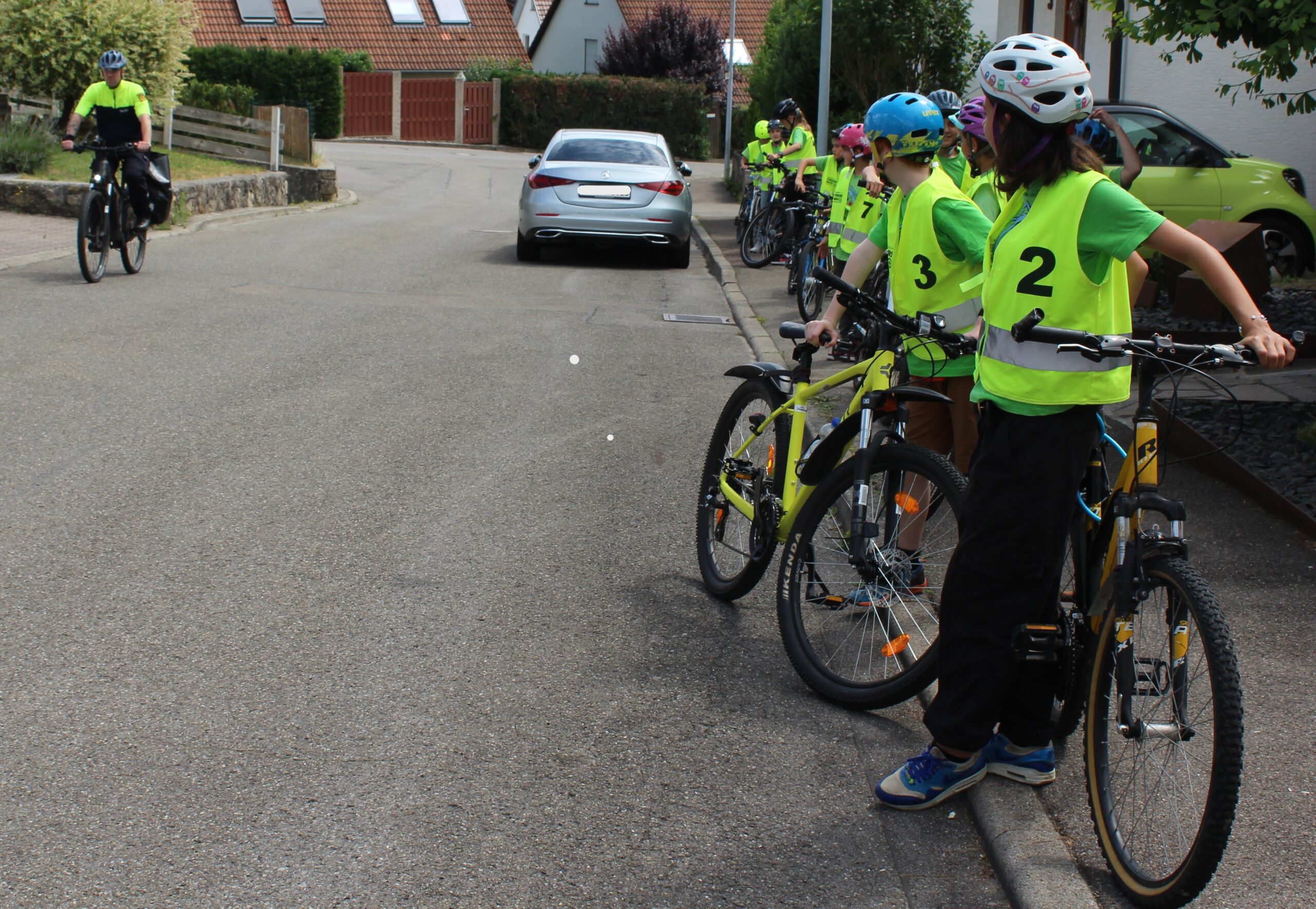 An einer Straße steht eine Klasse mit ihren Fahrrädern und Leuchtwesten und schaut auf einen Polizisten auf dem Fahrrad.