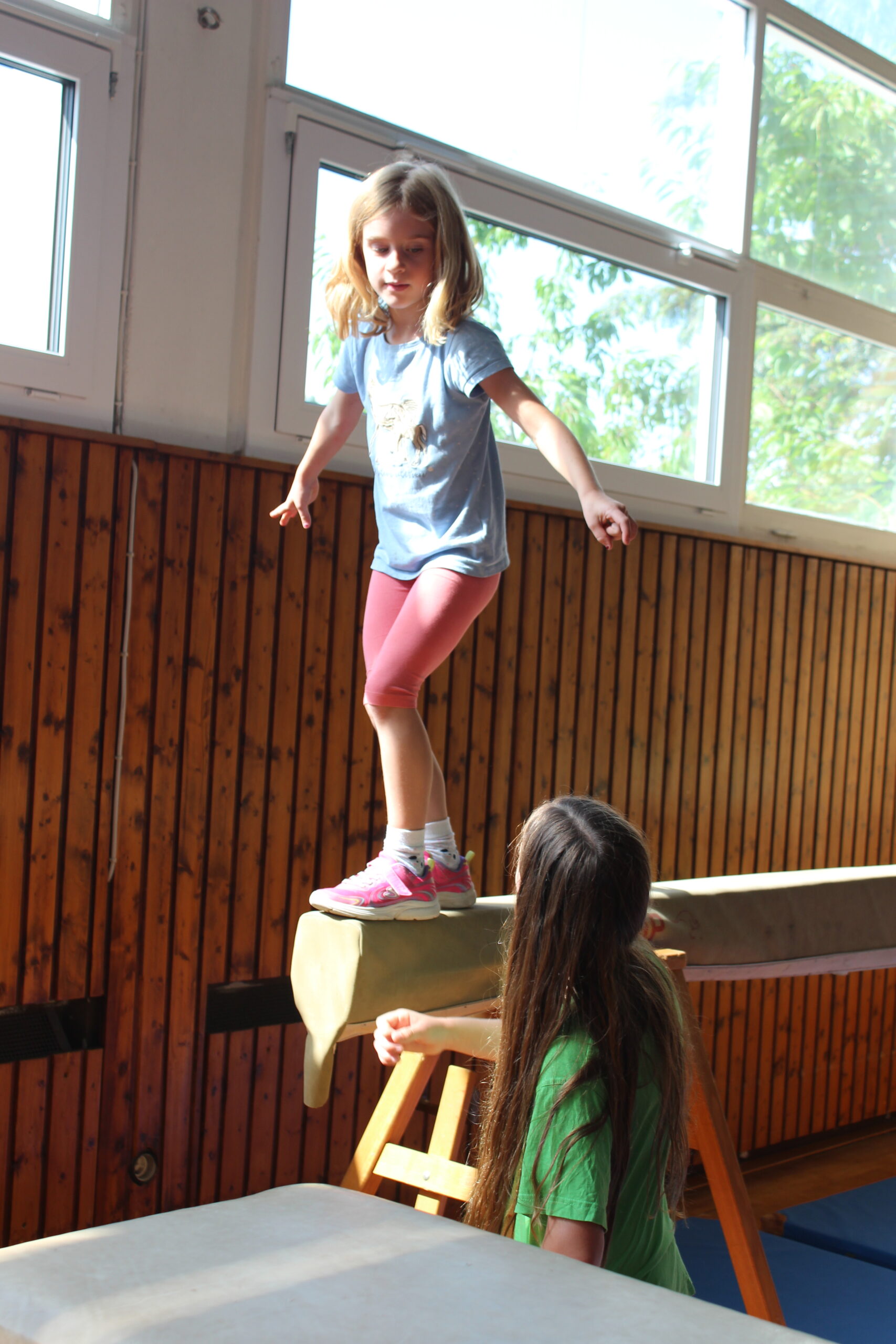 Ein Kindergartenkind balanciert auf einem Schwebebalken und ein Schulkind gibt Hilfestellung.