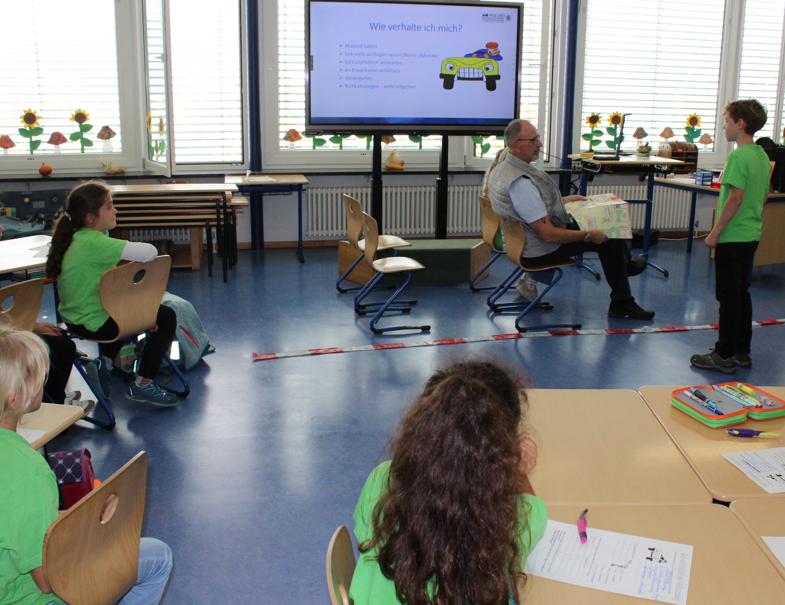 In einem Klassenzimmer ist eine Spielsituation aufgebaut, die ein Auto darstellt, dessen Beifahrer ein Kind auf der gespielten Straße anspricht.