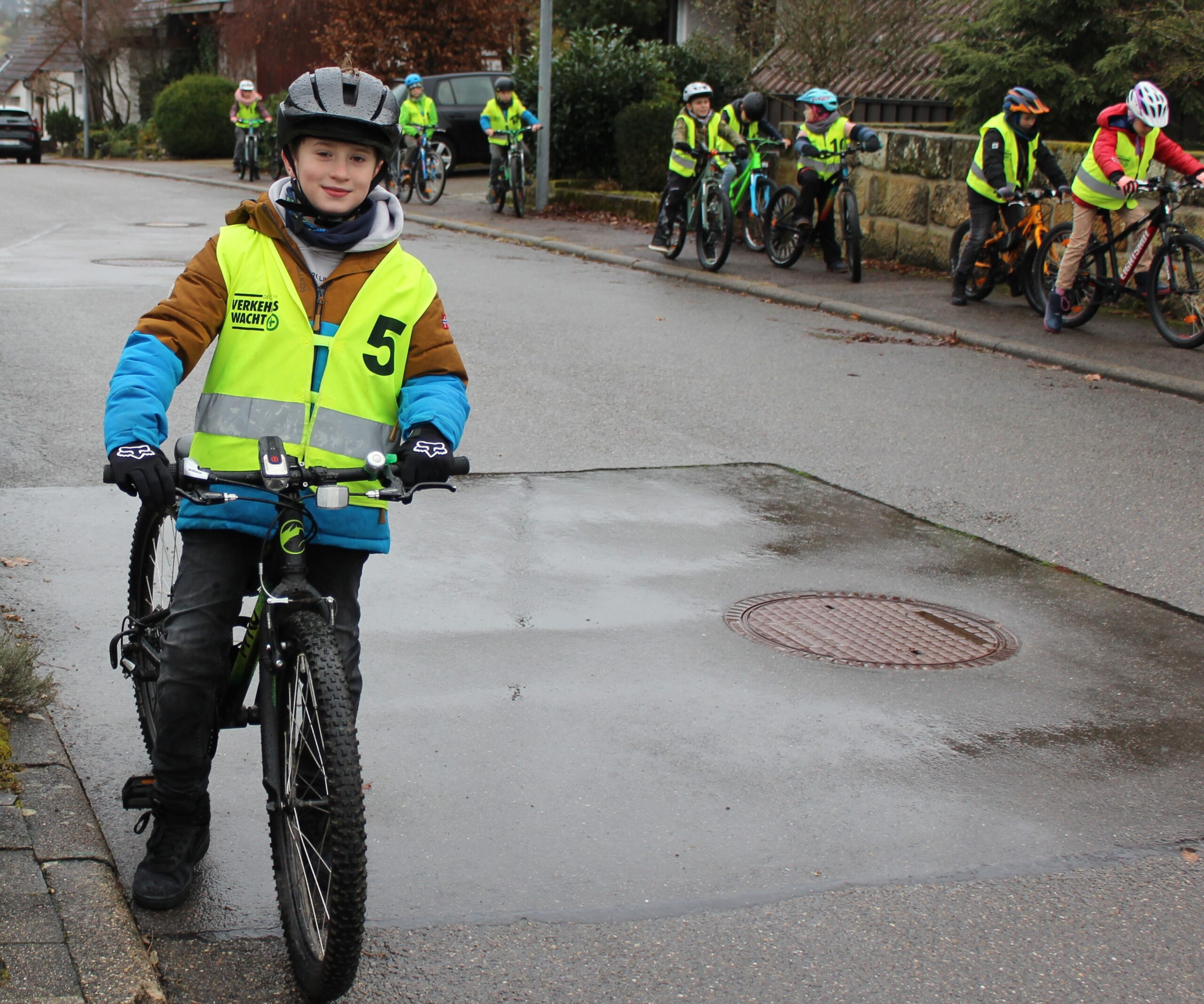 Im Vordergrund steht ein Kind bereit zum Losfahren und im Hintergrund sieht man andere Kinder auf dem Gehweg Fahrrad fahren.