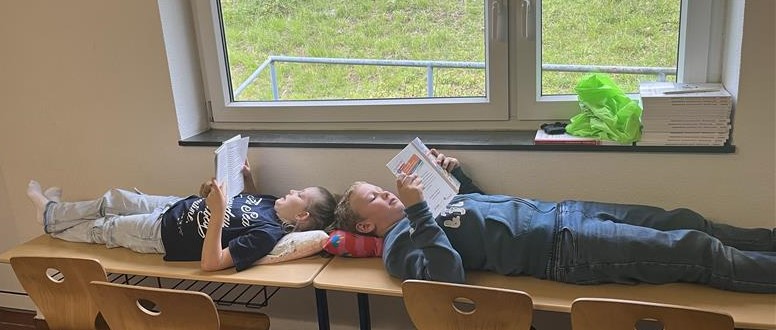 Zwei Jungen liegen auf Tischen in einem Schulgang und lesen.