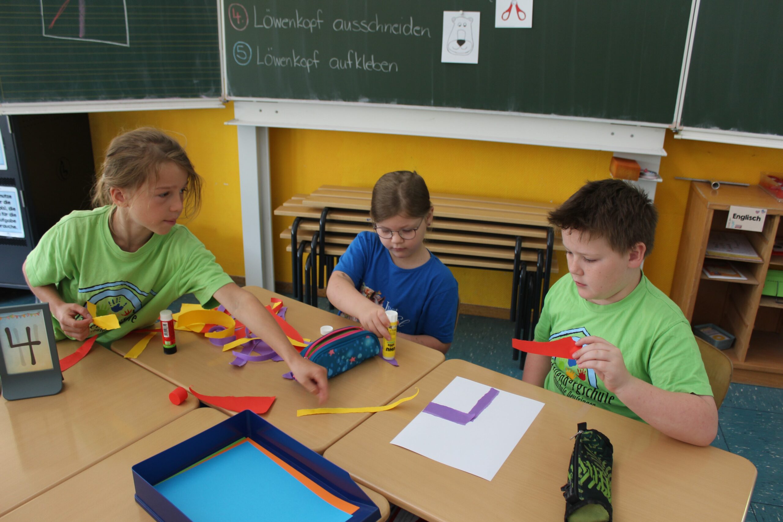 Kinder basteln an einem Schultisch. Zwei tragen ein Schul-T-Shirt und das dritte Kind ist ein Kindergartenkind.