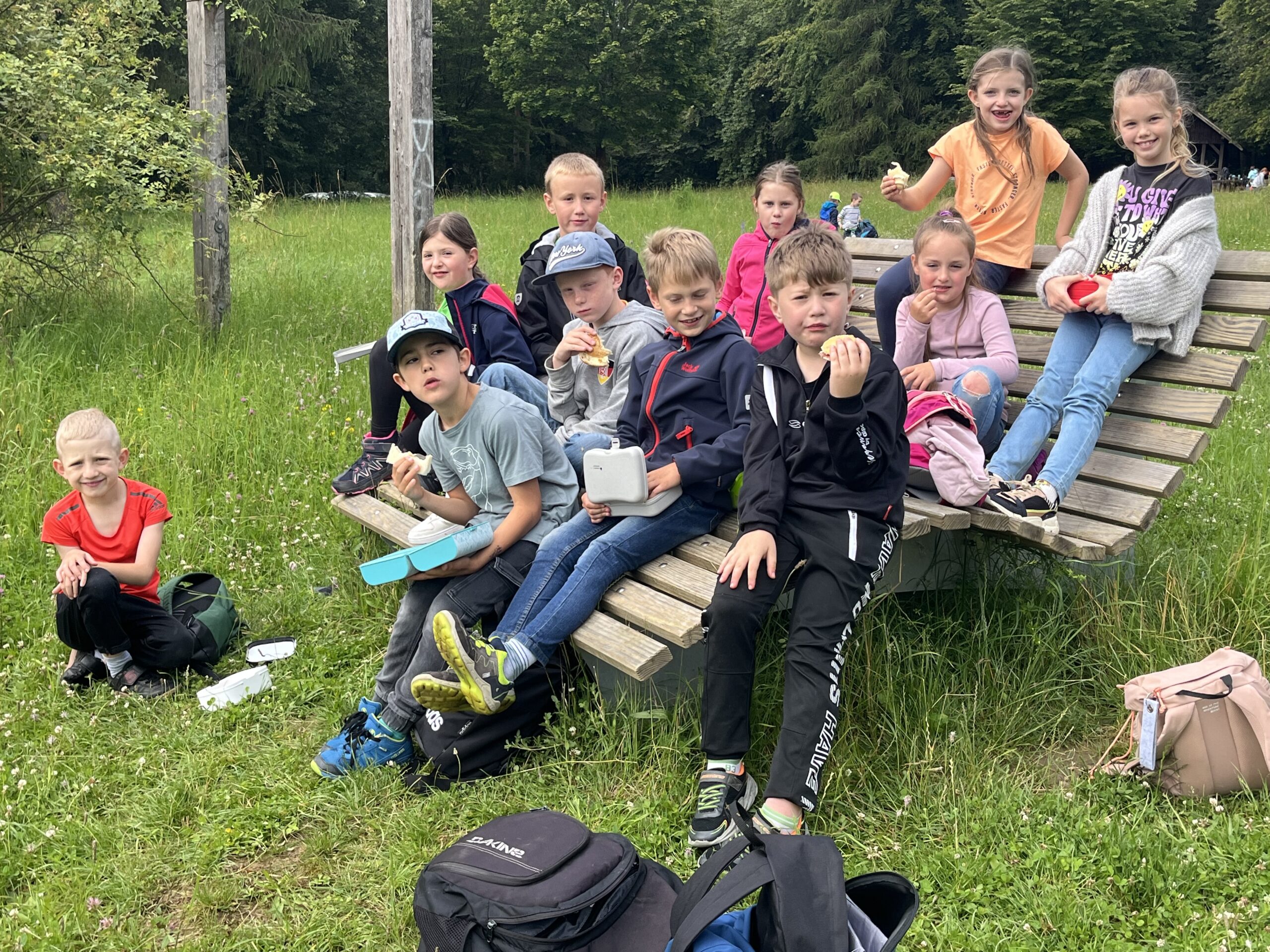 Eine Schulklasse sitzt auf einer großen Bank bei ihrem Wandertag und vespert.