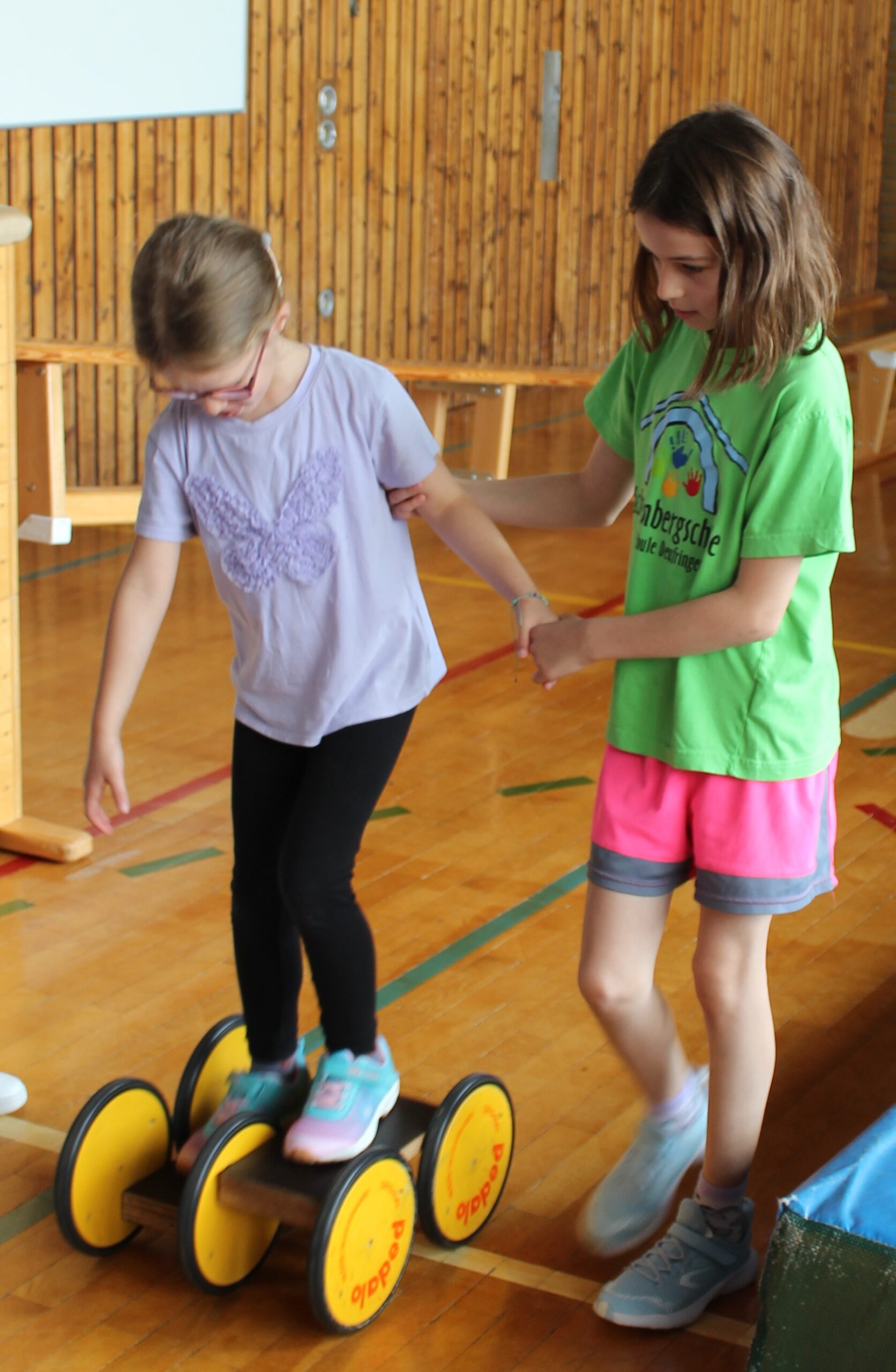 Ein Schulmädchen hält ein Kindergartenkind an der Hand, das gerade auf einem Pedalo fährt.