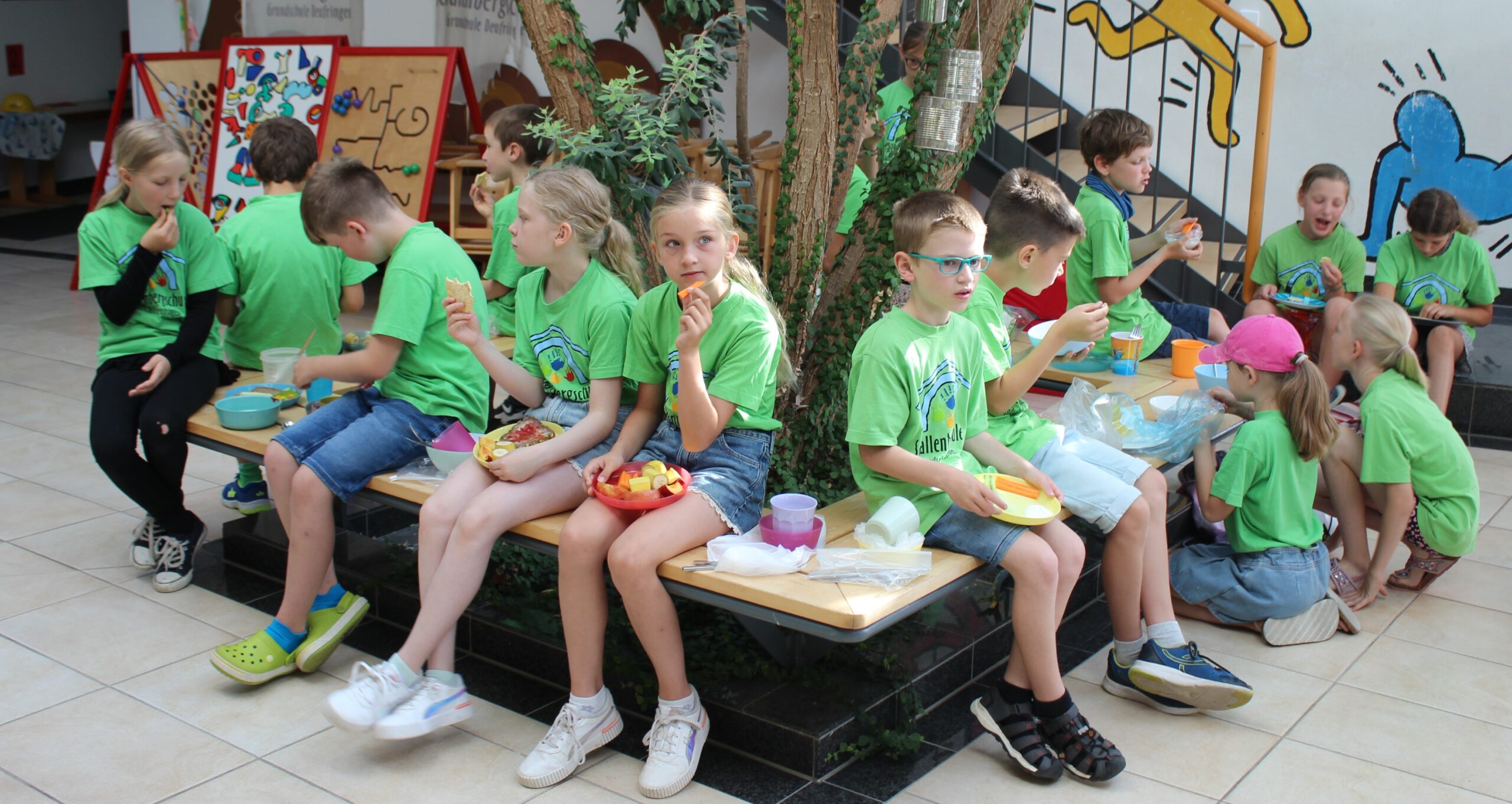 Kinder essen auf einer Bank um einen Baum in der Aula.