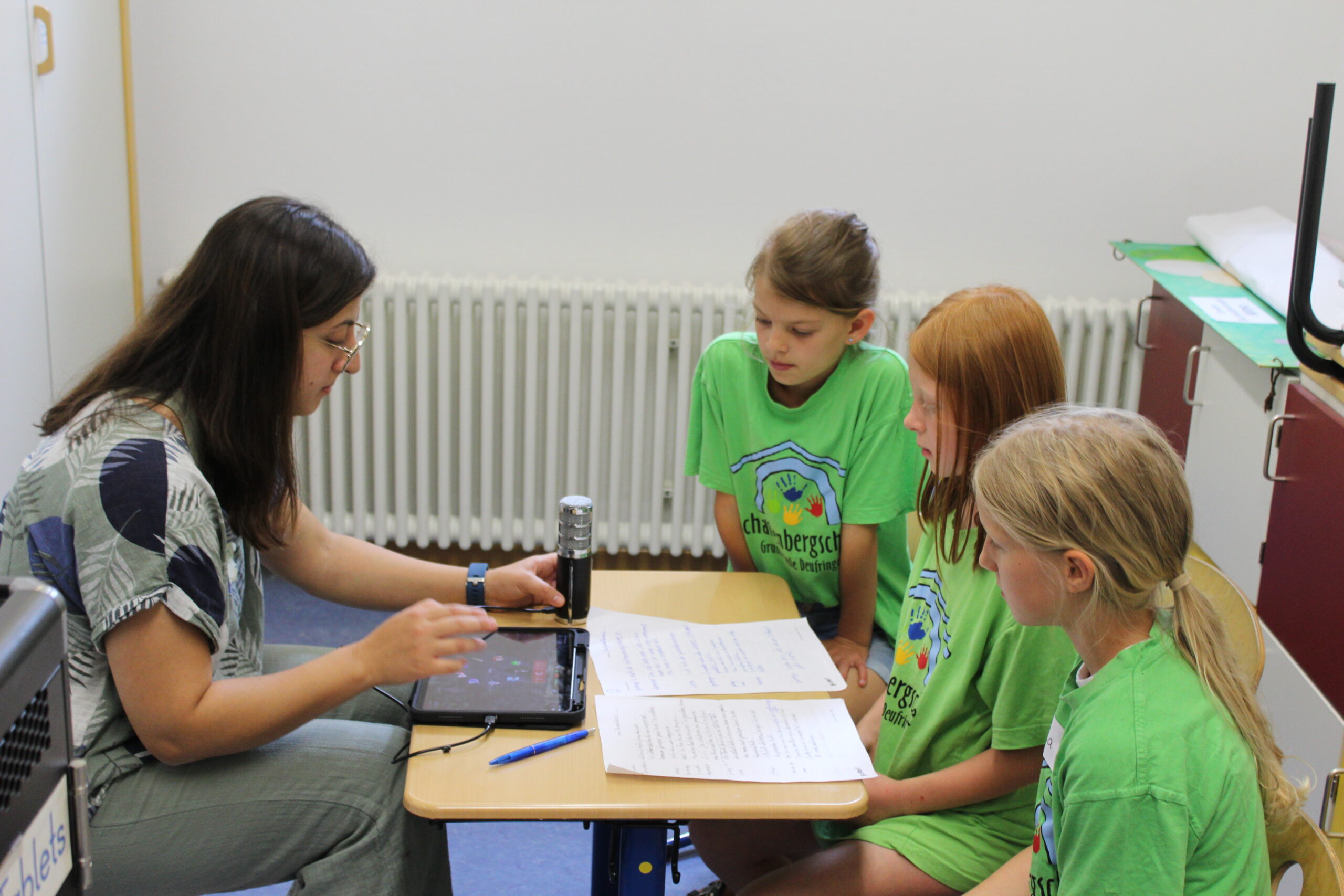 Frau und drei Kinder sitzen sich an einem Tisch gegenüber, auf dem Tisch steht ein Mikrofon, ein Tablet und Texte; sie nehmen etwas auf