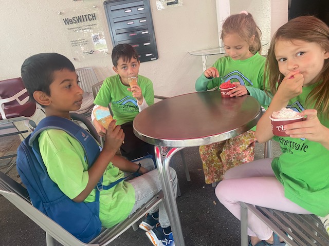 vier Kinder sitzen um einen runden Tisch herum, sie halten ein Eis in der Hand und essen es