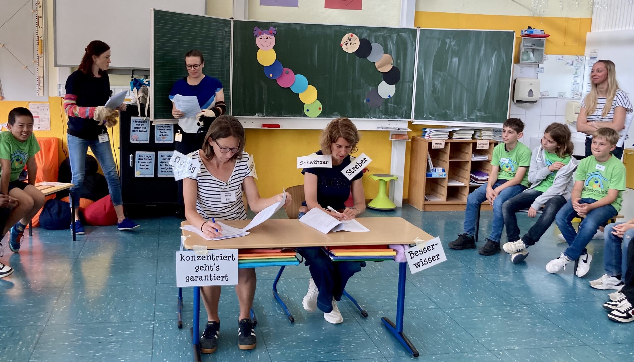 zwei Frauen an einem Tisch, lesen etwas, am Tisch kleben Karten mit den Aufschriften "konzenriert geht's garantiert" und "Besserwisser", im Hintergrund zwei weitere Frauen, lesen etwas vor