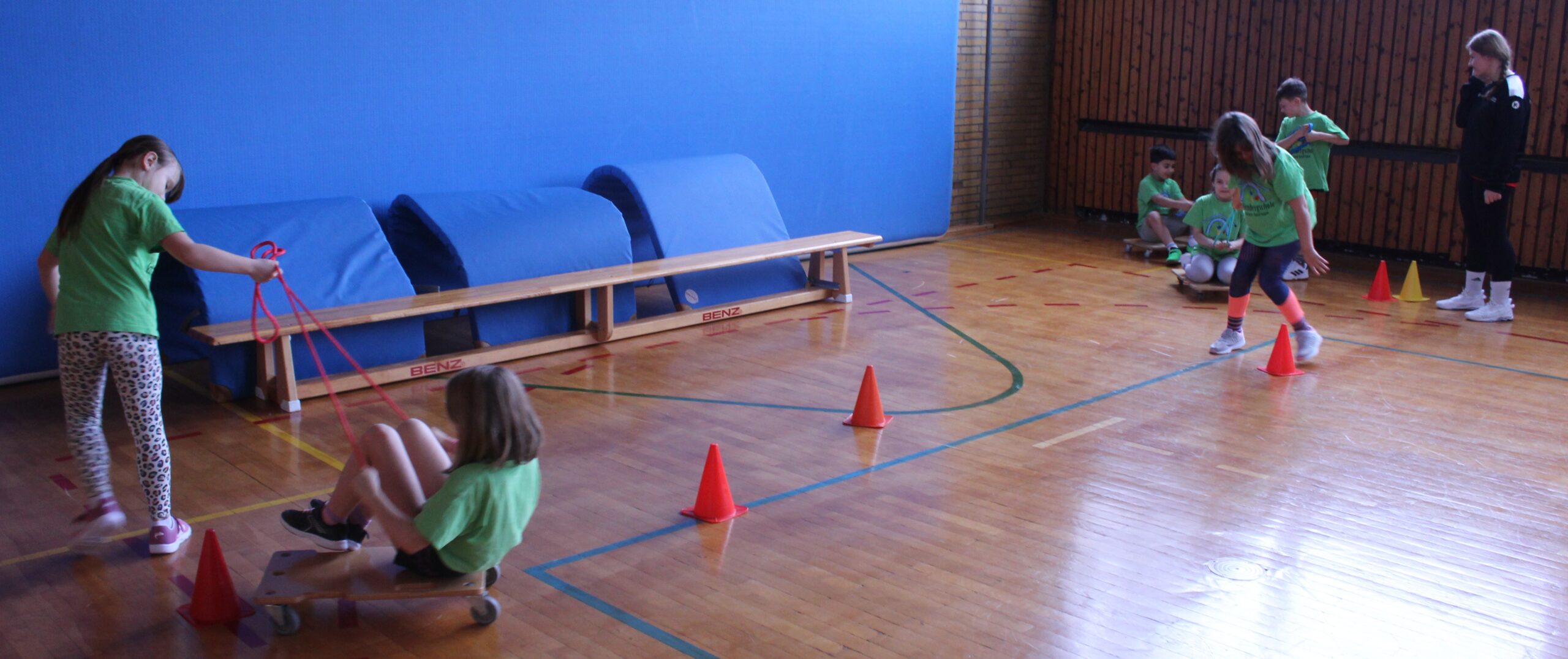 Hier zieht ein Kind ein anderes auf dem Rollbrett durch einen Mattentunnel und durch Slalomhütchen.