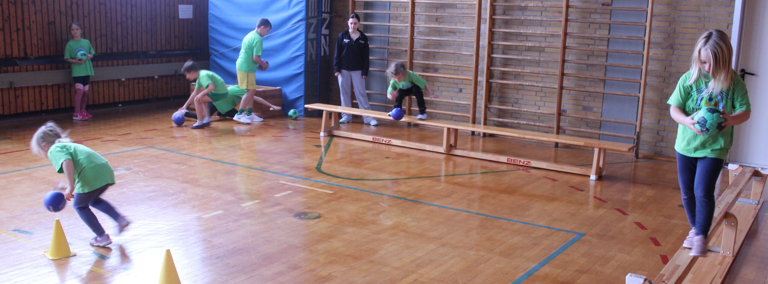 Kinder laufen auf einer Bank und prellen dabei daneben mit einem Ball.