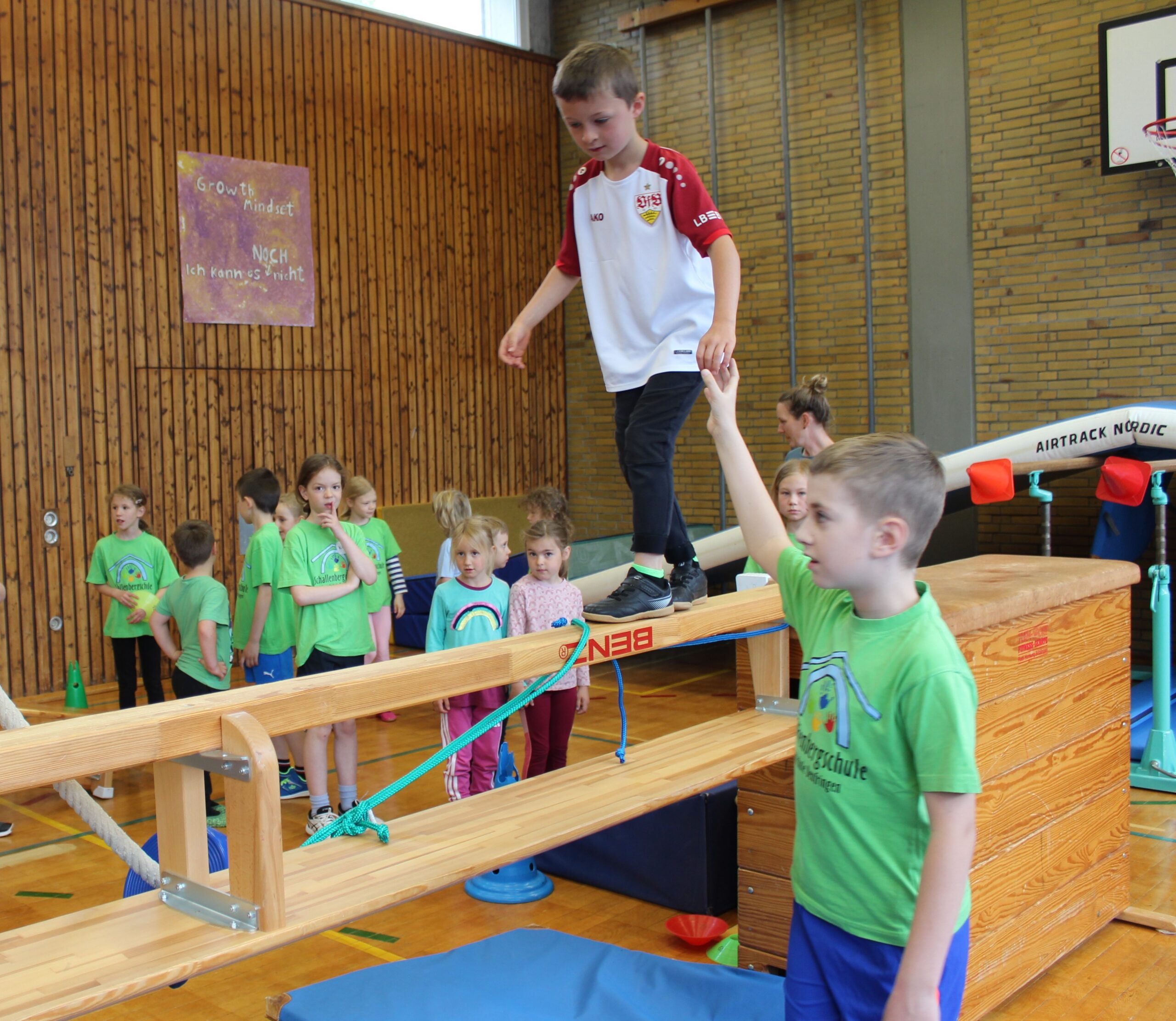 Ein Drittklässler gibt einem Kindergartenkind Hilfestellung beim Balancieren auf einer umgedrehten Bank, die zwischen zwei hohe Kasten eingebaut ist.